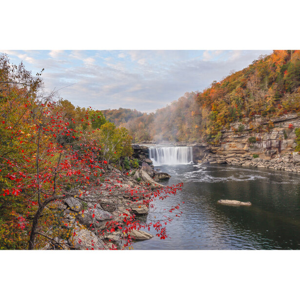 Waterfall flowing over rocky ledges with autumn foliage in foreground and forested canyon walls.