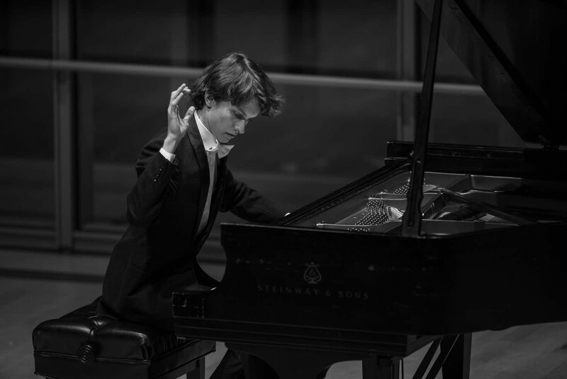 Pianist in formal attire seated at a grand piano, hand raised to head in a contemplative gesture.