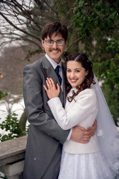 Newlyweds posing together outdoors, groom in gray suit and bride in white dress with veil.