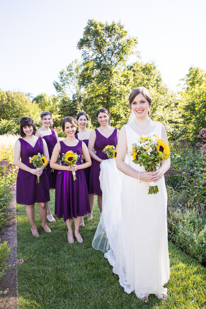 Bride in white gown with bridesmaids in purple dresses holding yellow bouquets outdoors.