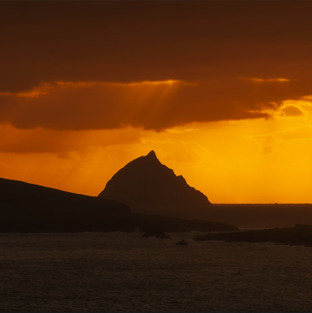 Silhouetted rocky island against golden sunset sky with dramatic clouds and water in foreground.