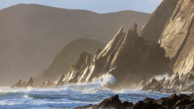 Dramatic coastal cliffs with columnar basalt formations rise above crashing waves and black rocks.