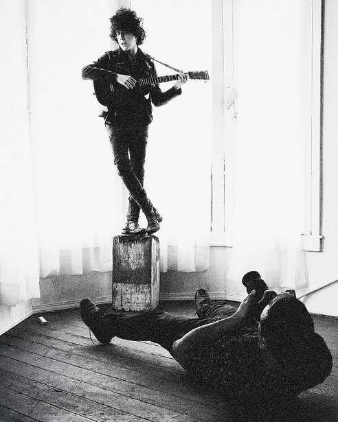 Musician standing on an amplifier playing electric guitar while another person lies on the floor below.