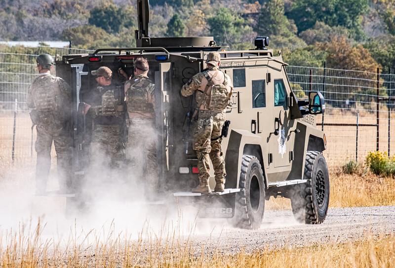 Military personnel in camouflage uniforms conduct dust control exercises around an armored tactical vehicle on a dirt road.