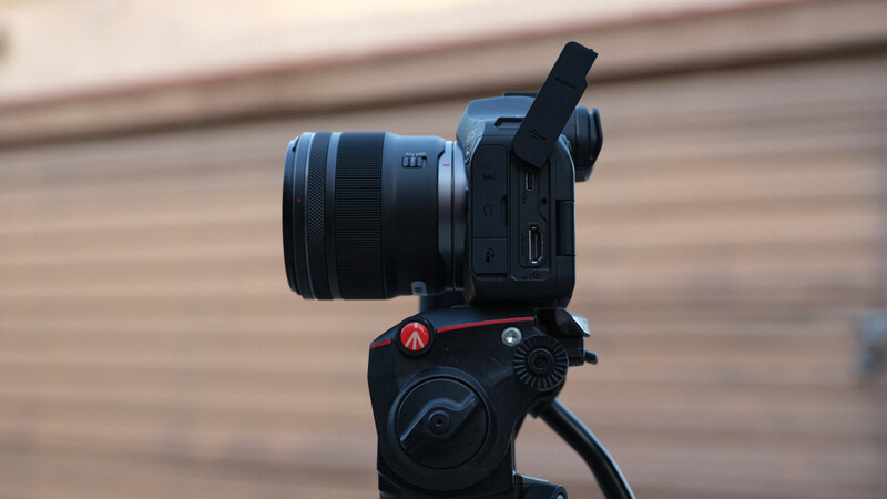 Compact cinema camera with attached lens mounted on a fluid head tripod, viewed from the side against a wooden interior background.