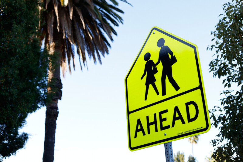 Bright yellow pedestrian crossing warning sign with silhouettes of an adult and child, flanked by palm and ivy foliage.