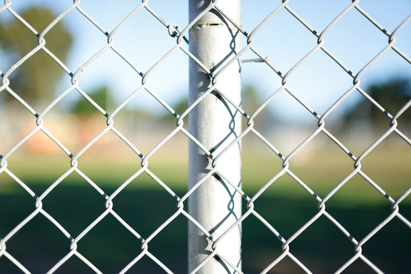 Chain-link fence with white metal post in sharp focus, blurred green field and trees in background.