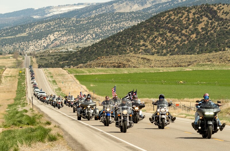 Motorcycle riders lead a long line of vehicles down a rural highway with forested hills in the background.