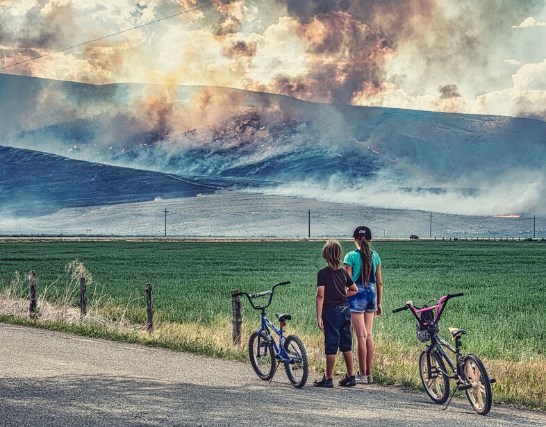 Three cyclists standing on a road beside green fields with a massive wildfire and smoke billowing across a distant mountain.