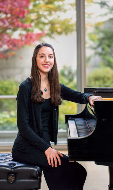 Woman in black blazer seated at grand piano with flowering vines in background.