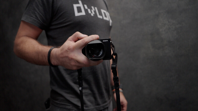 Photographer holding a compact camera with a leather strap against his chest, wearing a dark branded t-shirt.