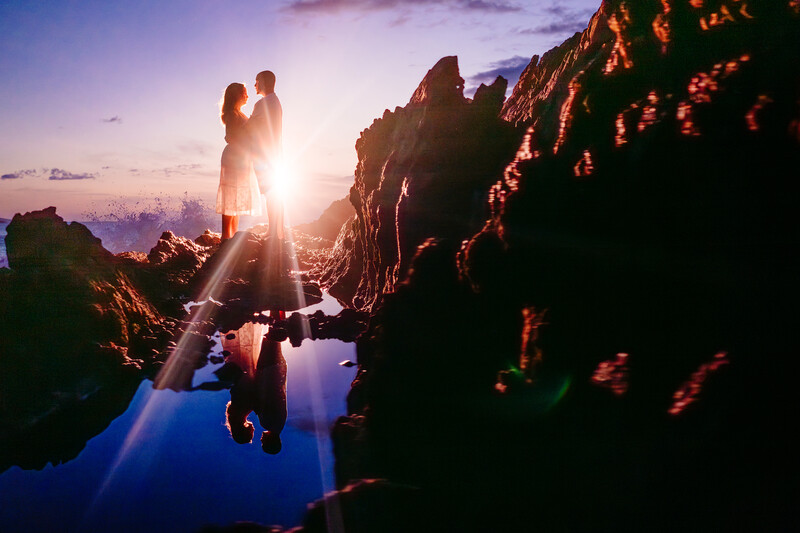 Couple standing on rocky outcrop silhouetted against sunset sky with photographer in foreground.