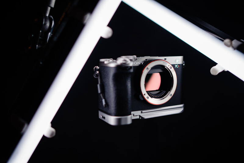 Mirrorless camera body with exposed sensor mounted on ring light stand against black background.