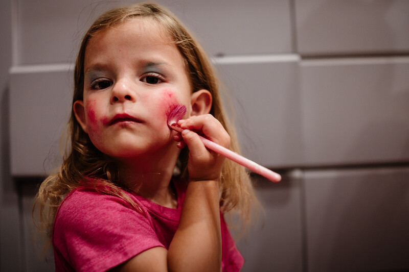 Young girl applying pink makeup to her cheek with a brush, looking at camera.