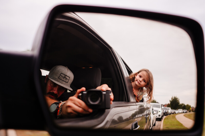 Reflection in side mirror showing photographer holding camera while shooting from moving vehicle window.