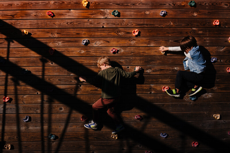 Overhead view of two people on a wooden climbing wall with colorful handholds casting sharp shadows.
