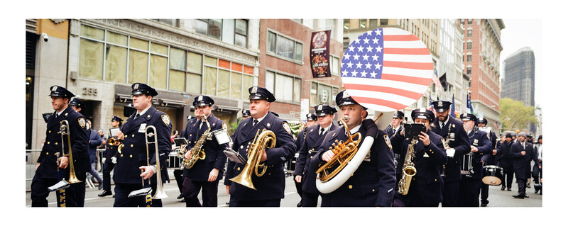 Police marching band in formation on urban street with American flag overhead.