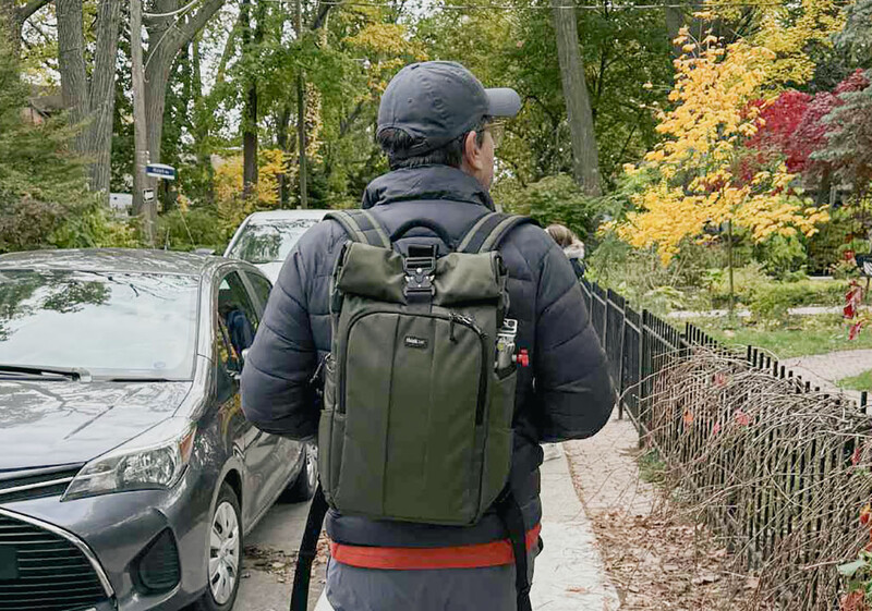 Photographer wearing olive backpack and black jacket stands on residential street during autumn.