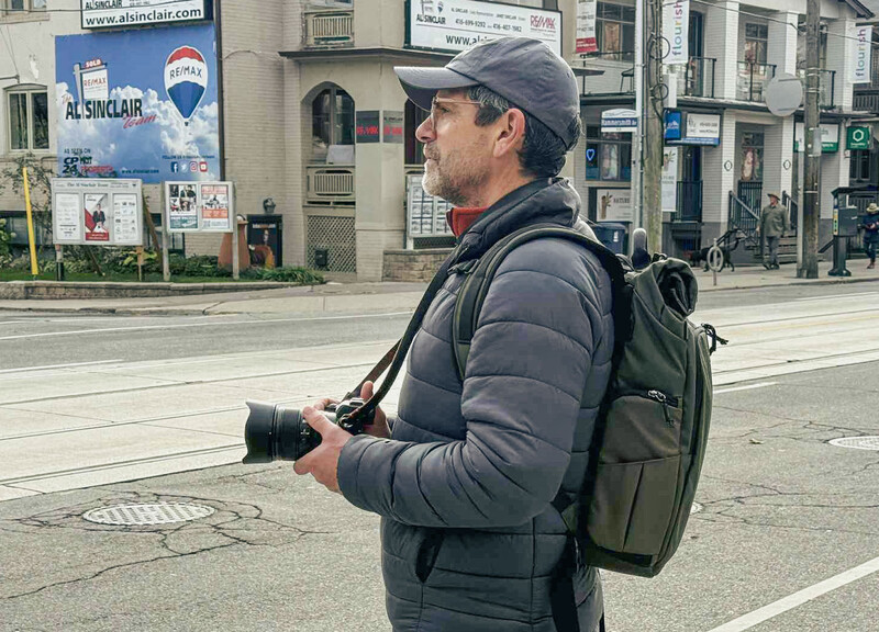 Photographer holding a DSLR camera with telephoto lens, wearing a gray puffer jacket and backpack on an urban street.