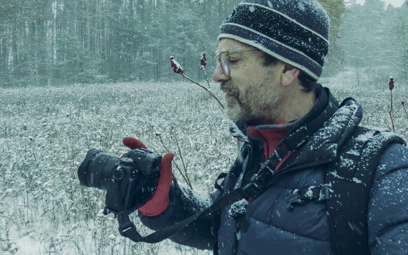 Photographer holding a camera in a snowy forest landscape with mountains in background.