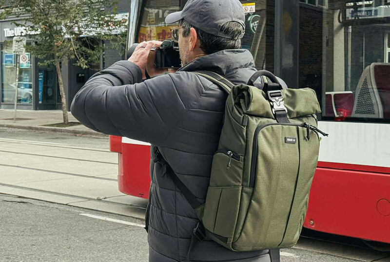 Photographer with camera and olive backpack shooting from a street with a red bus in the background.