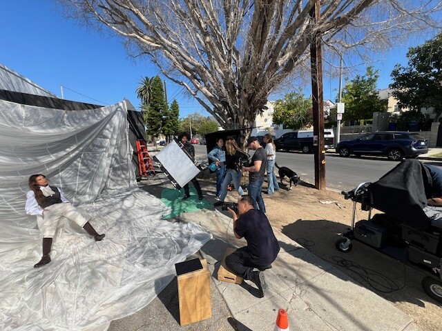 Film crew setting up a scene on a sunny street with a large reflector and shade cloth positioned near a leafless tree.