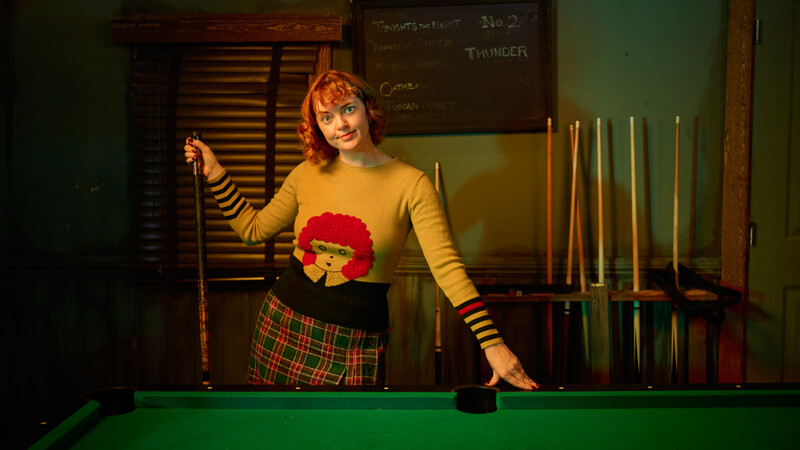 Woman in yellow striped sweater and plaid skirt holds pool cue at billiards table.