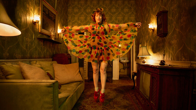 Woman wearing a colorful polka dot cape strikes a pose in a vintage-style bedroom with warm gold wallpaper and layered lighting.