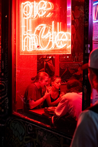 Red neon sign glowing above people seated at a table in a dimly lit venue.