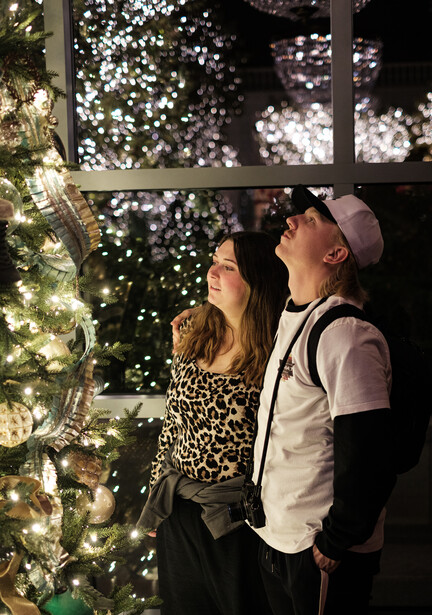 Two people gazing upward at an illuminated Christmas tree display visible through a window at night.