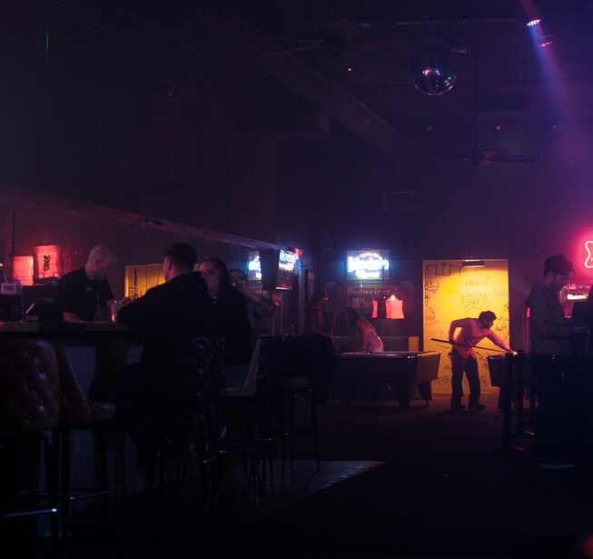 Dark indoor venue with silhouetted figures and warm amber and cool blue neon lighting throughout the space.