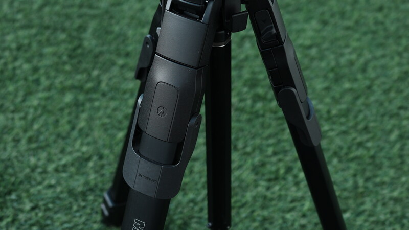 Close-up of a black tripod head with adjustment knobs and control buttons against a green textured background.