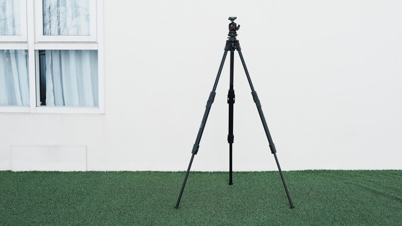 Professional camera tripod with three extended legs on grass in front of a white building.