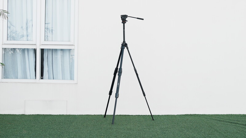 Black tripod with fluid head mounted on a grassy outdoor surface against a white building.