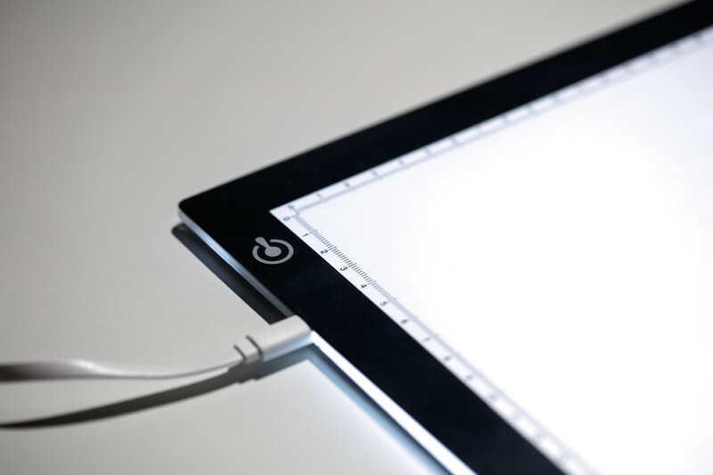 Close-up of a light pad's corner showing the power button and connected USB cable against a neutral background.