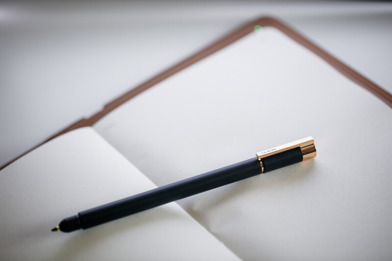 Black pen with gold trim resting on an open blank notebook with brown leather cover.