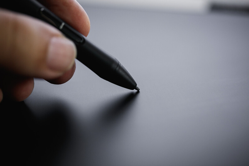 Close-up of a stylus pen tip held between fingers against a blurred gray surface.