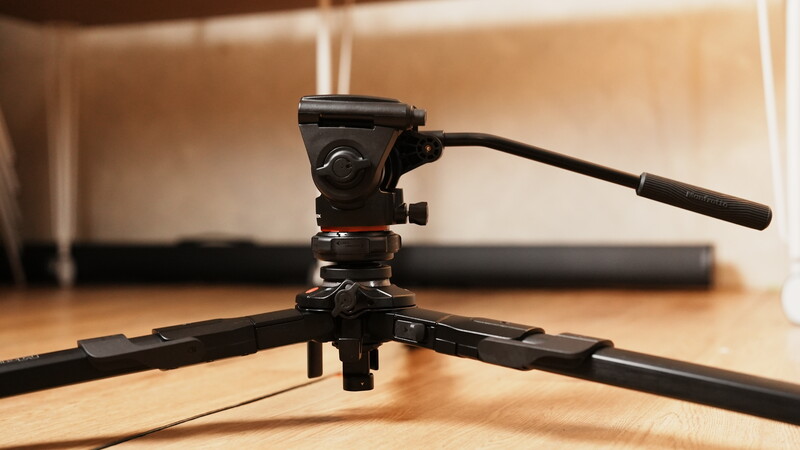 Fluid head video tripod with pan handle mounted on black spreader legs on wooden surface.