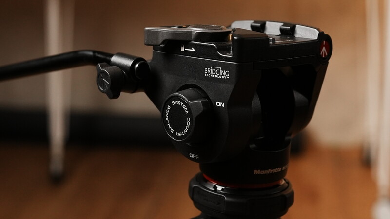 Black fluid head video tripod with pan-tilt handle and control knobs mounted on wooden surface.