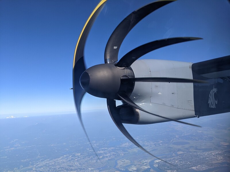 Turboprop aircraft engine in flight with propeller blades visible against blue sky and landscape below.