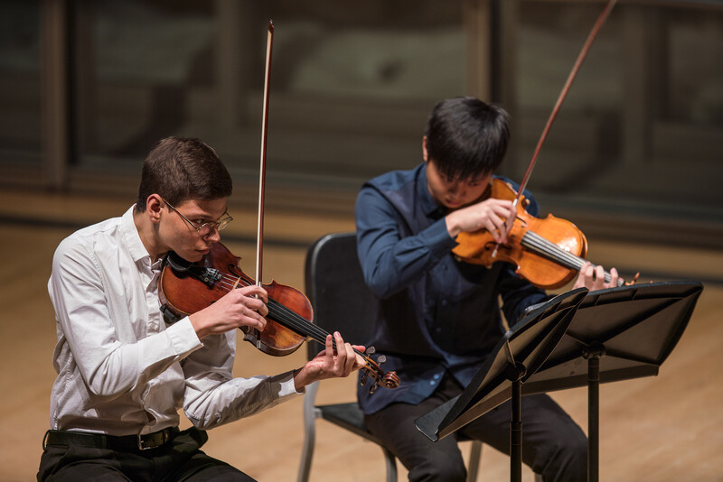 Two violinists performing together on stage with sheet music on a stand.