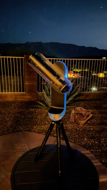 Telescope on tripod pointing at night sky with mountains and lit railing in background.