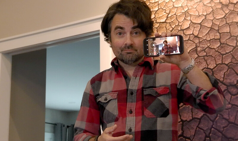Man in red plaid shirt holding smartphone displaying video call with another person.