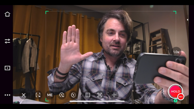 Screenshot of a video call showing a man in a plaid shirt holding up his hand while looking at a mobile device.