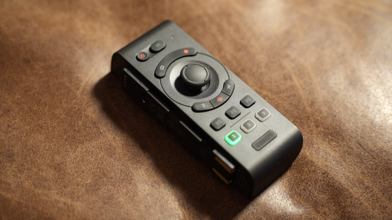 Black wireless remote control with rotary dial and tactile buttons on textured brown surface.