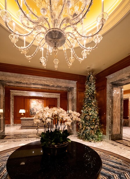 Luxurious hotel lobby with ornate crystal chandelier, marble flooring, and holiday decorations including decorated tree.