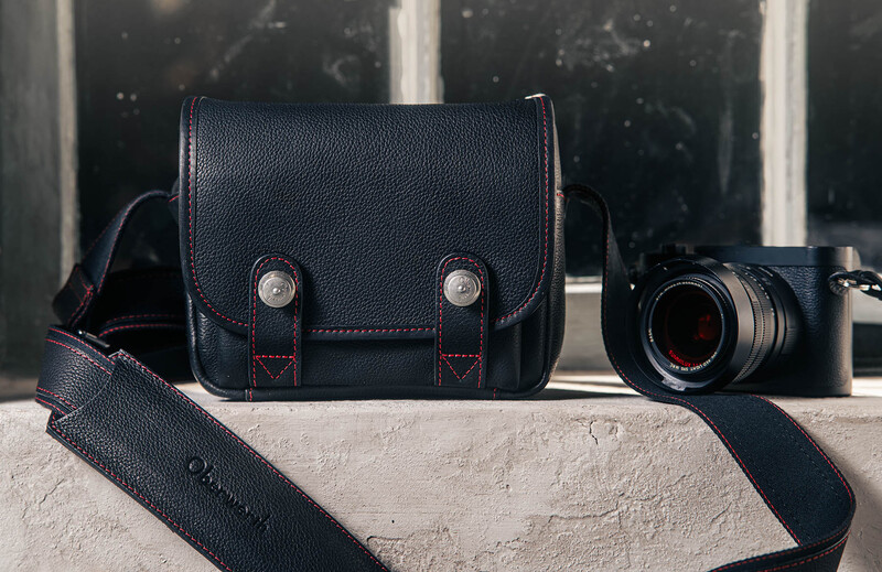 Black leather camera bag with red stitching and brass hardware displayed next to a vintage rangefinder camera.