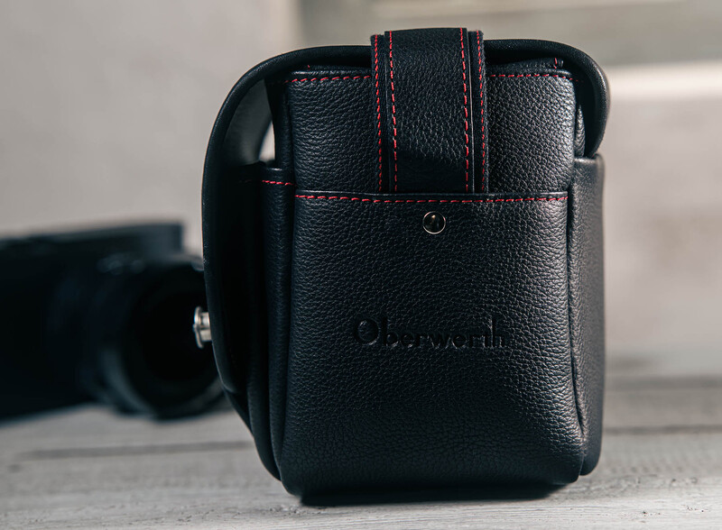 Black leather camera bag with red stitching and shoulder strap displayed on concrete surface.