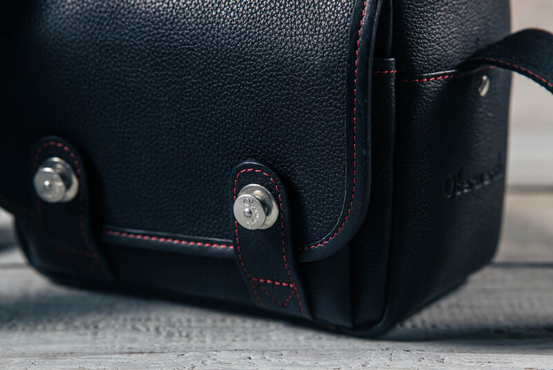 Close-up of black leather camera bag with red stitching and metal hardware details.