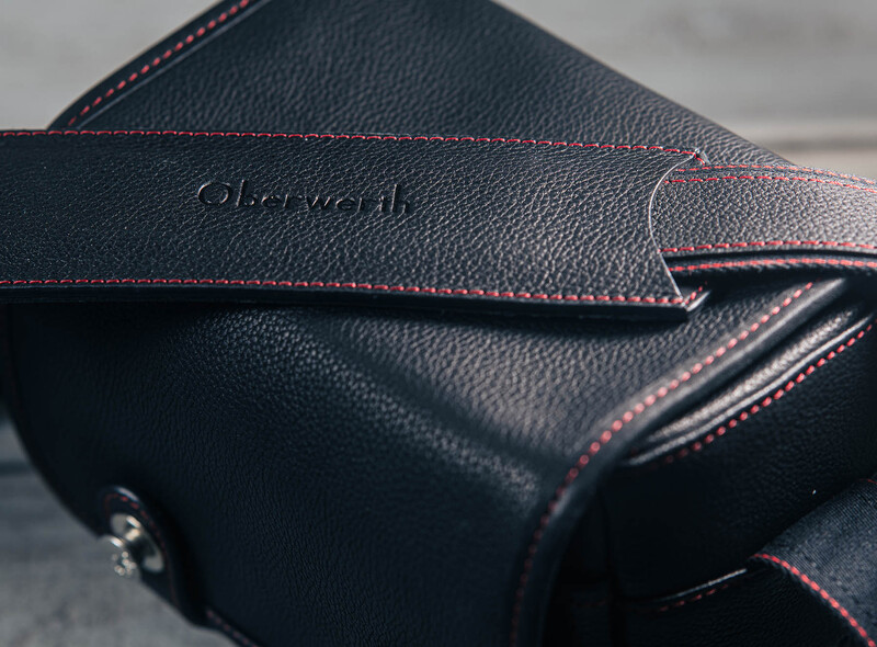 Black leather camera bag with red stitching and embossed branding detail.